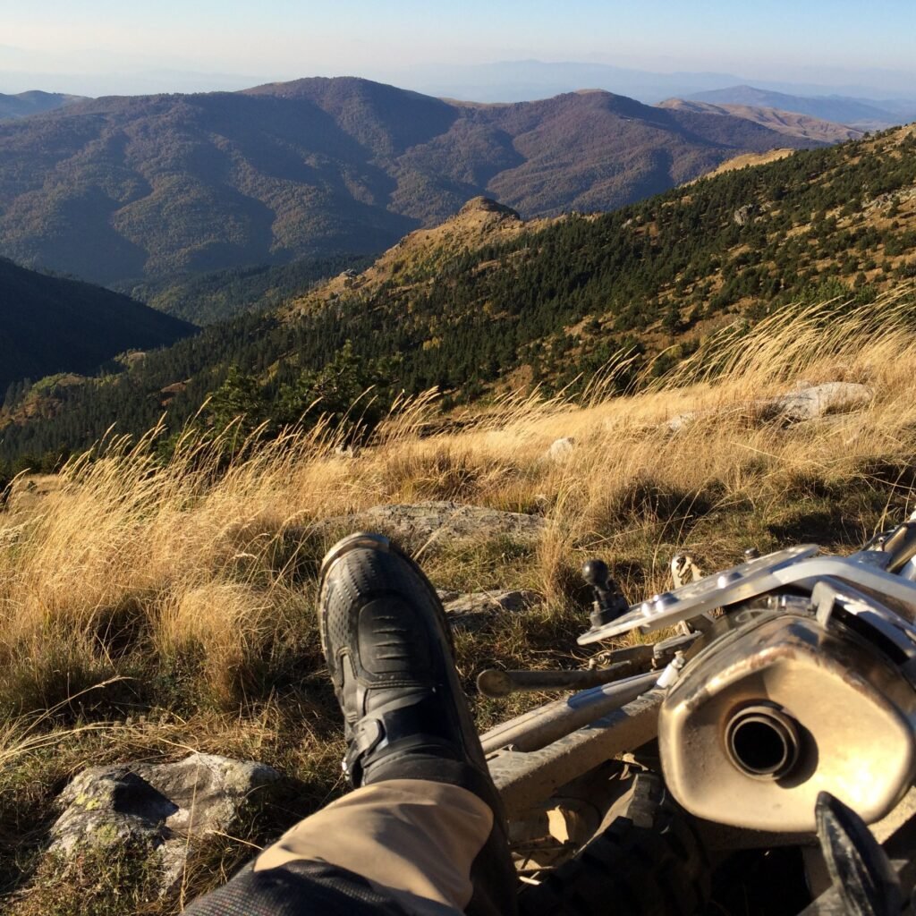 Mountain landscape with a close-up of an off-road motorcycle and the rider’s enduro boot, symbolizing the start of an off-road journey.